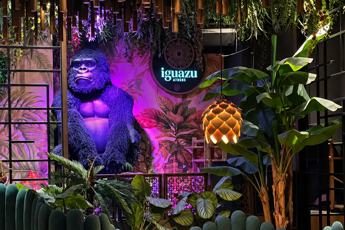 The giant gorilla under violet light at night, beneath the iguazu Athens neon sign, with a pinecone lamp and banana palms beside him
