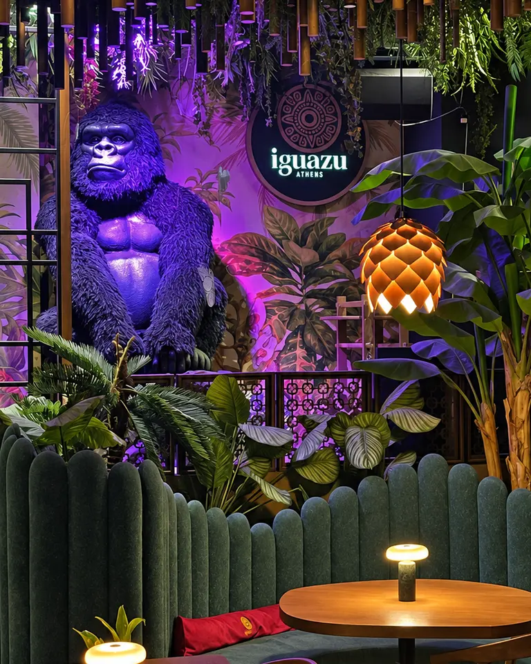 The giant gorilla lit violet above the planters at night at Iguazu Athens, seen across the dining room from a table